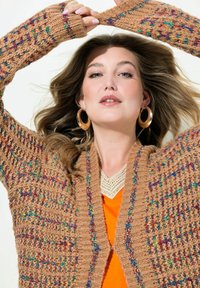 Cardigan en tricot marron avec des motifs colorés, à l'avant ouvert, en tissu texturé et avec des poignets côtelés. Se superpose à un haut orange et présente des accents en dentelle.