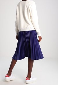 Sweatshirt de couleur crème avec poignets côtelés associé à une jupe midi plissée marine. Baskets blanches avec des accents rouges au talon.