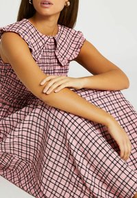 Rutig rosa klänning med rynkad krage och smockad livstycke. Tyget har en krinklad struktur med svarta rutmönster.