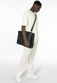 Schwarze Lederhandtasche mit strukturierten Oberflächen, doppelten Griffen und verstellbarem Schultergurt, kombiniert mit einem hellen Outfit und weißen Schuhen.