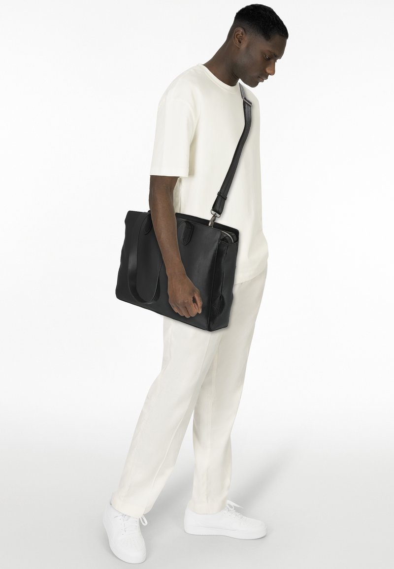 Schwarze Lederhandtasche mit strukturierten Oberflächen, doppelten Griffen und verstellbarem Schultergurt, kombiniert mit einem hellen Outfit und weißen Schuhen.
