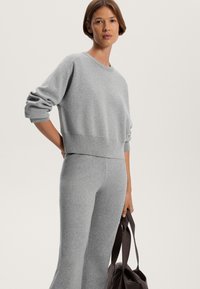 Grauer, kurz geschnittener Sweatshirt mit langen Ärmeln und gerippten Bündchen, kombiniert mit passenden, enganliegenden grauen Hosen, gehalten mit einer dunkelbraunen Tragetasche.