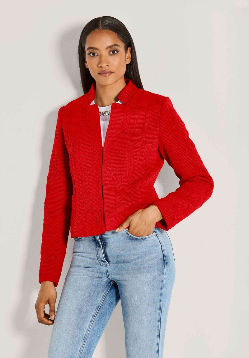 Rote, strukturierte, kurze Jacke mit Kragen und langen Ärmeln, mit einem geometrischen Muster und Fronttaschen, getragen über hellblauen Jeans.