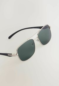 Zonnebril met een zilver metalen frame en groene lenzen. Voorzien van zwarte plastic pootjes en een unieke geometrische lensvorm.