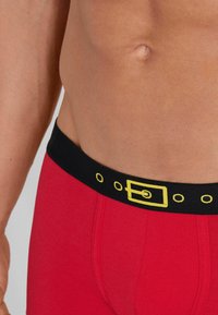 Červené bavlněné boxerky s černým páskem, který má design žluté přezky na opasku a kruhové detaily, zdůrazňující přiléhavý střih.