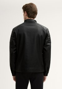 Giacca in pelle nera con accenti imbottiti sulle spalle, colletto alto e texture liscia. La vista posteriore mostra una vestibilità sartoriale.