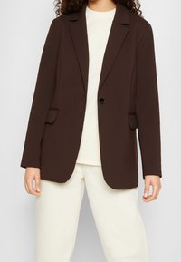Blazer marrone in tessuto liscio con un solo bottone, reverse a scollo aperto e due tasche frontali. Indossato sopra una maglietta chiara e pantaloni crema.