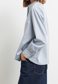 Personne portant une chemise bleu clair à manches longues boutonnée et un jean bleu foncé avec poches, debout de côté avec une main dans la poche.