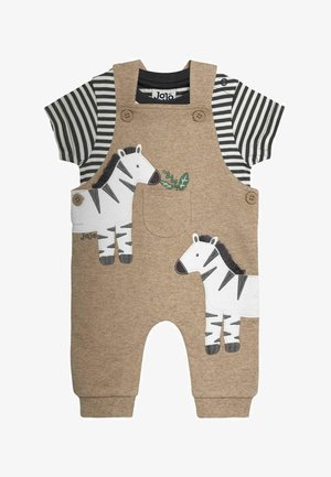 Beżowe ogrodniczki dla małego dziecka z aplikacjami zebry, noszone na czarno-białej koszulce w paski z krótkim rękawem, z zapinanymi na guziki paskami i małą kieszonką na piersi.