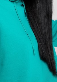 Sweat à capuche turquoise avec un cordon de serrage et un tissu texturé ; cheveux noirs longs et raides partiellement visibles sur la droite.