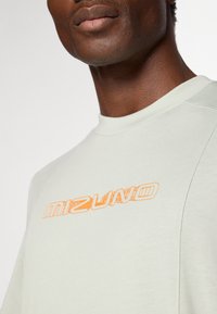 Svetlozelené bavlnené tričko s okrúhlym výstrihom, s veľkým oranžovým logom "Mizuno" na hrudi a textúrovanými detailmi látky.