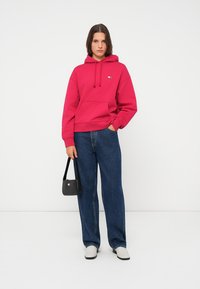 Lys rosa hettegenser med frontlomme og hette, sammen med løse blå jeans og hvite slip-on sko. Svart veske med logo i hånden.
