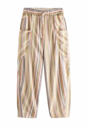 Kvinde står indenfor ved en trædør iført en cremefarvet top med sorte blomsterærmer og brede, stribede bukser.