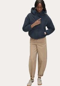 Marineblå pufferjakke med hætte, med lynlås fortil, elastiske manchetter og to forlommer, parret med beige brede bukser og sneakers.