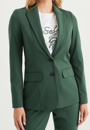 Vrouw in een nauwsluitend donkergroen colbert met twee zwarte knopen, klepzakken en bijpassende broek, gedragen over een wit T-shirt met grafisch ontwerp.