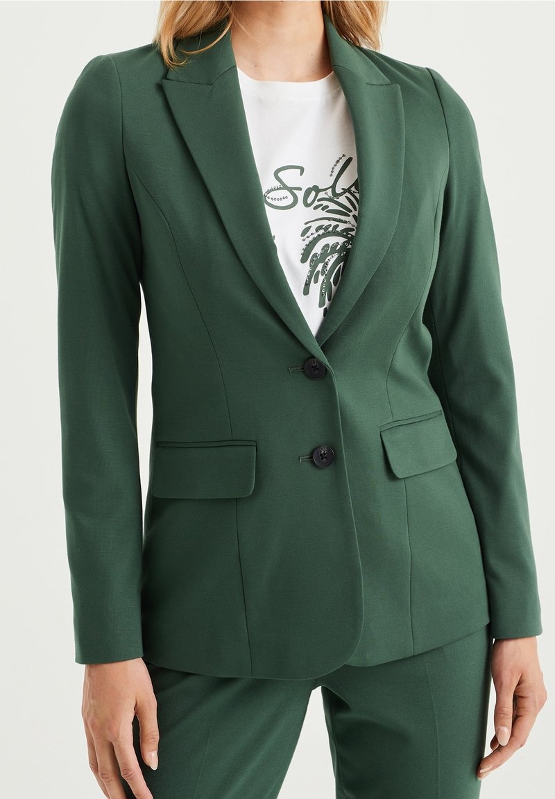 Femme portant un blazer vert foncé cintré avec deux boutons noirs, des poches à rabat, et un pantalon assorti, sur un tee-shirt blanc imprimé.