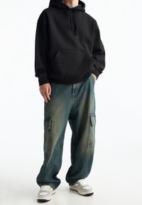 Svart huvtröja med framficka, kombinerad med löst sittande, urtvättade blå cargobyxor med sidofickor och vita sneakers.