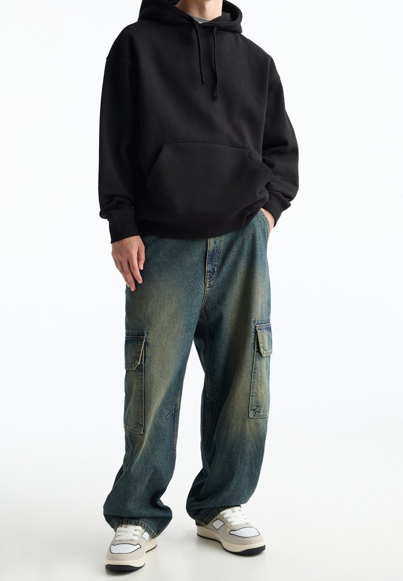 Svart huvtröja med framficka, kombinerad med löst sittande, urtvättade blå cargobyxor med sidofickor och vita sneakers.