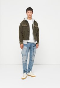 Olijfgroene denimjack met zilveren knopen, grijze hoodie eronder, gecombineerd met lichtblauwe cargo jeans en beige sneakers.