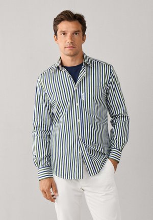 Hombre con camisa de rayas verticales en azul, verde y blanco sobre una camiseta azul marino y pantalones blancos, de pie frente a un fondo liso.