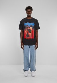 Sort T-shirt med grafisk tryk i retro-stil. Modellen har lys blå brede jeans og hvide sneakers på.
