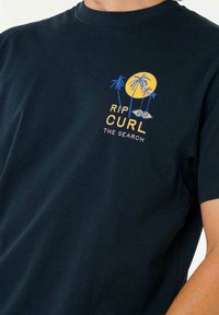 Navy blue cotton T-shirt featuring a graphic print with yellow sun, blue palm trees, and text "RIP CURL THE SEARCH" in vibrant colors.