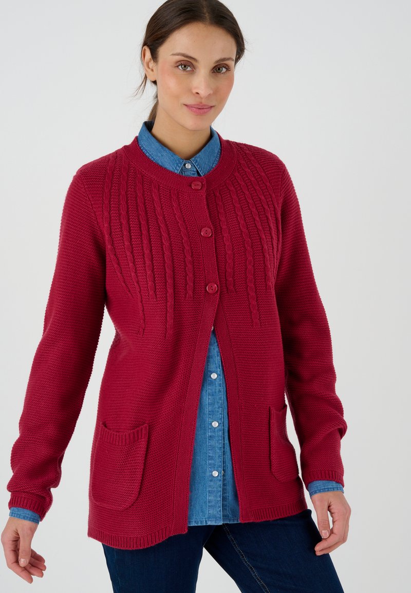 Cárdigan rojo de punto con un patrón texturizado, que presenta un cuello redondo, dos bolsillos delanteros y cierre de botones, superpuesto sobre una camisa de denim.