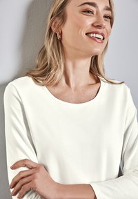 Camicia bianca a maniche lunghe con scollatura ampia e morbido tessuto. Il modello indossa un orecchino a cerchio dorato, con uno sfondo neutro.