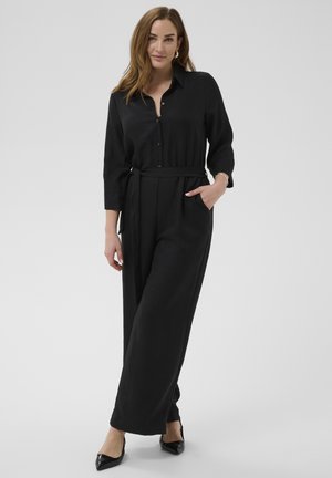 Kvinde poserer iført en sort jumpsuit med knapper, lange ærmer, taljebånd og sorte spidse hæle mod en ensfarvet baggrund.