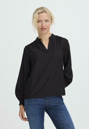 Schwarze Bluse mit langen Ärmeln, V-Ausschnitt und gerafften Schulternahtdetails, kombiniert mit blauen Jeans. Weicher Stoff mit feiner Textur.
