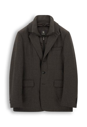 Homme portant une veste brune superposée, une chemise bleu clair à col, un jean foncé et des baskets marron clair, debout devant un fond uni.