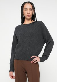 Γκρι υπερμεγέθη πλεκτή μπλούζα με ριμπ μανσέτες και τελείωμα. Μαλακή υφή, φαρδιά λαιμόκοψη και άνετη εφαρμογή, συνδυασμένη με καφέ παντελόνια.