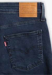 Jean en denim bleu foncé avec une poche arrière et un patch en cuir marron clair de la marque Levi's sur la ceinture.