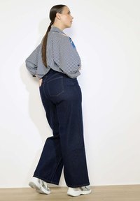 Pruhovaná oversized košile a tmavě modré široké džíny. Džíny mají zadní kapsy a jsou doplněny bílými a šedými teniskami.