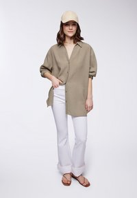 Camicia verde oliva con bottoni, dal taglio morbido, abbinata a pantaloni bianchi e sandali marroni. La camicia presenta un tessuto texturizzato e maniche arrotolate.
