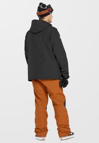 Zwarte waterdichte jas met capuchon, gecombineerd met oranje sneeuwbroeken. Zwarte beanie met wit patroon en bruine skibril. Zwarte handschoenen.