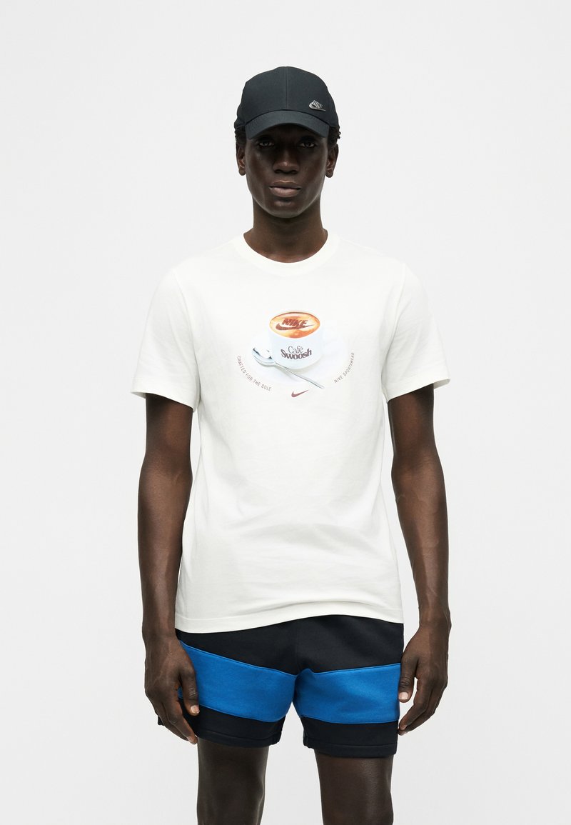 Giovane uomo che indossa una maglietta bianca Nike con grafica di una tazza di caffè, pantaloncini neri e blu e un cappellino nero Nike, in piedi contro uno sfondo bianco.