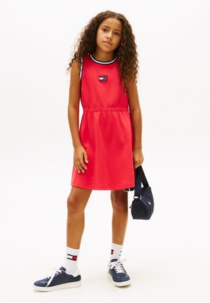 Mladé dievča s kučeravými vlasmi v červenom šatách bez rukávov, námornícke tenisky, biele ponožky, držiace malú námornícku tašku na jednoduchom pozadí.