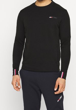 Mann trägt ein schwarzes Sweatshirt mit langen Ärmeln und passende schwarze Hose, beide mit kleinen Tommy Hilfiger-Logos und gestreiften Bündchen an den Handgelenken.