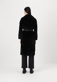 Abrigo de piel sintética negra con cuello, cintura ceñida con cinturón y mangas largas. Llevado sobre pantalones negros de pernera ancha y botines, visto desde atrás.