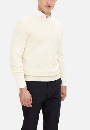 Maglione beige a trecce con orlo e polsini a coste, scollo rotondo, abbinato a pantaloni scuri, che mostrano un motivo zigzag testurizzato.