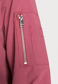 Detail rukávu maroon (bordó) bundy s vertikální stříbrnou zipovou kapsou a menší obdélníkovou nášivkou vedle ní.