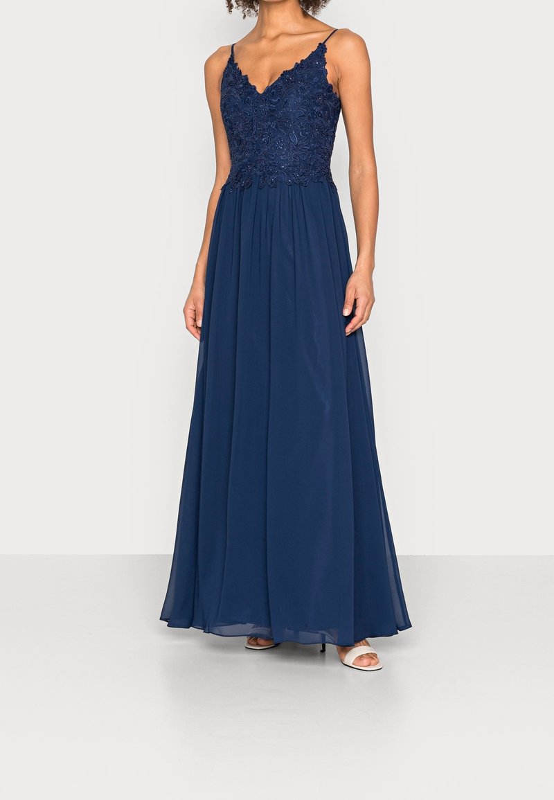 Femme portant une robe de soirée sans manches bleu marine avec un corsage en dentelle et une jupe fluide en chiffon, associée à des escarpins blancs à bouts ouverts.