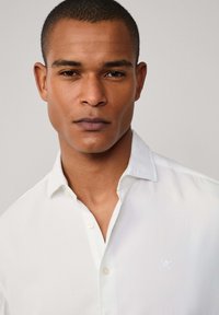 Close-up van een man met kort haar die een wit overhemd met knopen draagt, tegen een effen lichtgrijze achtergrond.