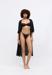 Schwarzer Morgenmantel mit Spitzenbesatz über einem Set aus schwarzer und burgunderfarbener Unterwäsche. Der BH hat ein halbtransparentes Design; der Tanga weist eine Spitzenakzentuierung auf.