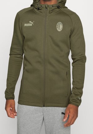 Mężczyzna w oliwkowo-zielonej bluzie zapinanej na suwak AC Milan z logo Puma oraz szarych spodniach dresowych z logo Puma, stojący na jednolitym tle.