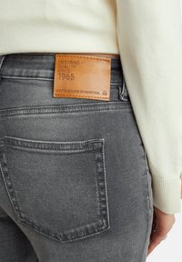 Šedé džíny z denimu s koženou nášivkou označenou "Trvalá kvalita od roku 1965", mají standardní kapsy a měkký, strukturovaný povrch.