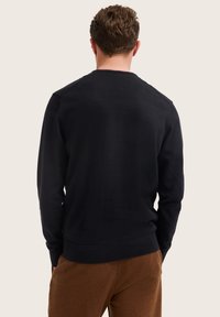 Mann, der mit dem Rücken zur Kamera steht und einen schwarzen langärmeligen Pullover sowie braune Hosen trägt, vor einem schlichten hellen Hintergrund.