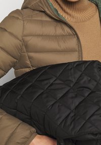 Ruskea puhallettava takki, jonka päällä on musta tikattu peitto, ja sen alla on ribbattu beige neule. Takissa on vetoketju ja vihreä vuori.