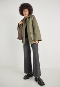 Geacă impermeabilă verde măslin cu închideri cu capse, două buzunare frontale, purtată cu blugi negri cu croială largă și loafers negri cu platformă.